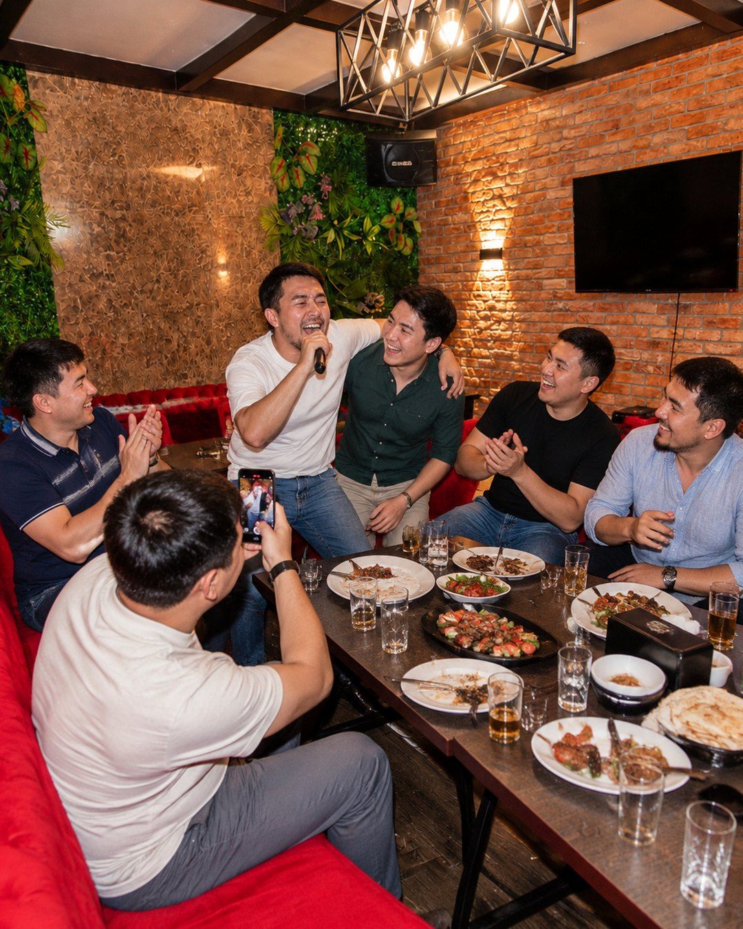 Bachelor party in an RMM room: the groom sings into a microphone with his arm around a friend, the others laugh and film on a phone